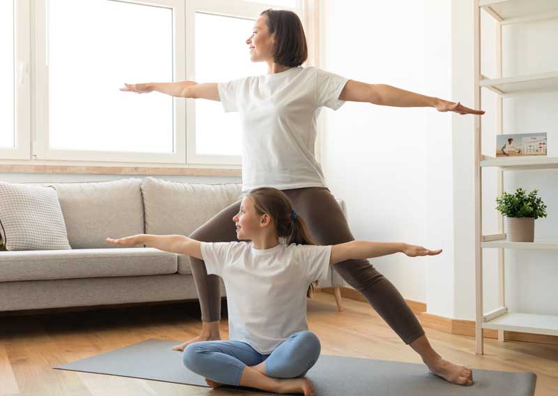 A woman and a young girl practicing yoga in a living room, both performing a warrior pose on yoga mats near a sofa and a bookshelf - corrective exercises A woman and a young girl practicing yoga in a living room, both performing a warrior pose on yoga mats near a sofa and a bookshelf - corrective exercises
