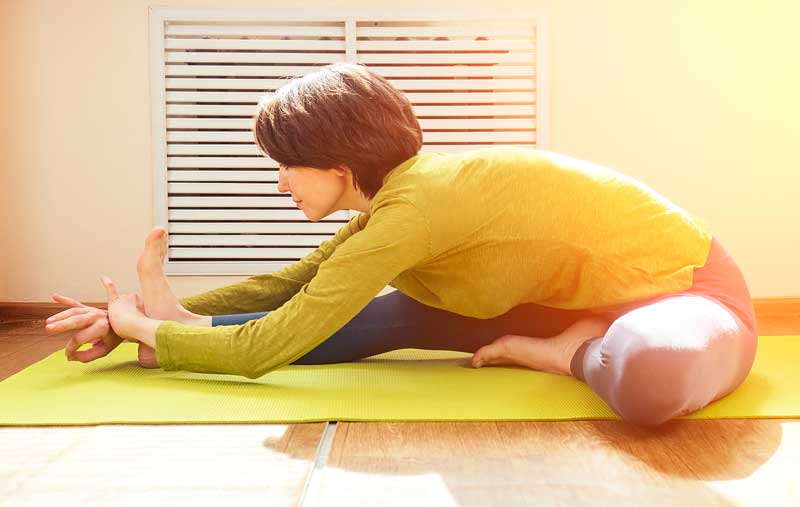A woman in a green top and leggings practicing yoga on a yellow mat, performing a seated forward bend in a sunlit room - extremity adjusting