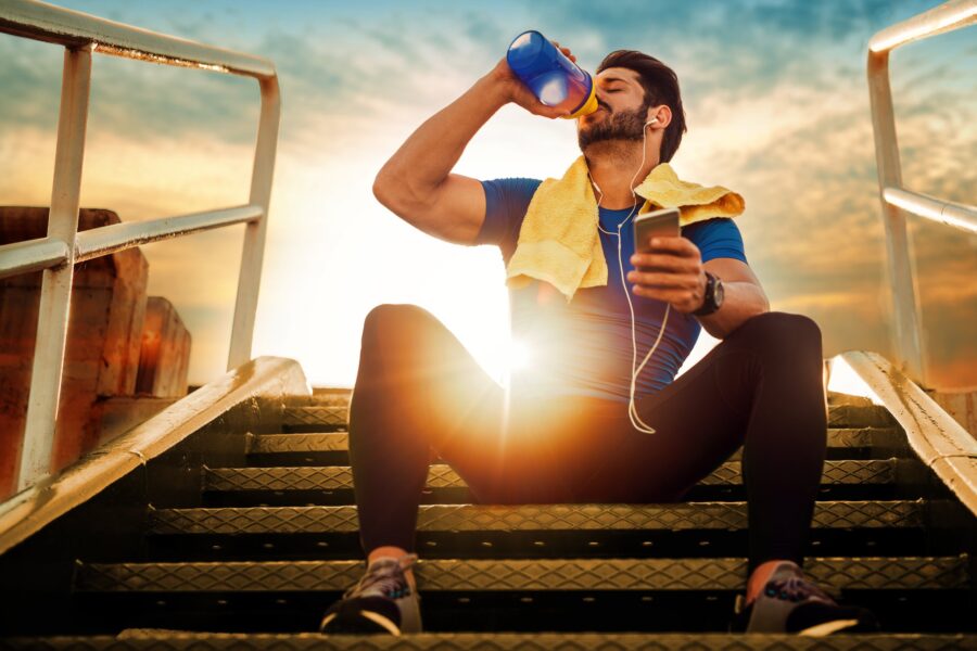 A fit man sitting on outdoor steps drinking water after a workout, holding a phone with headphones and towel, post-corrective exercise session - Corrective Exercises