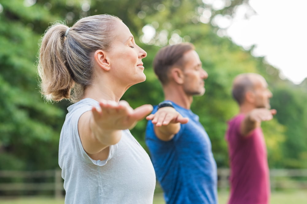 A group of adults performing yoga outdoors in a serene park, standing in a balanced pose with arms extended, focusing on mindfulness - corrective exercises A group of adults performing yoga outdoors in a serene park, standing in a balanced pose with arms extended, focusing on mindfulness - corrective exercises