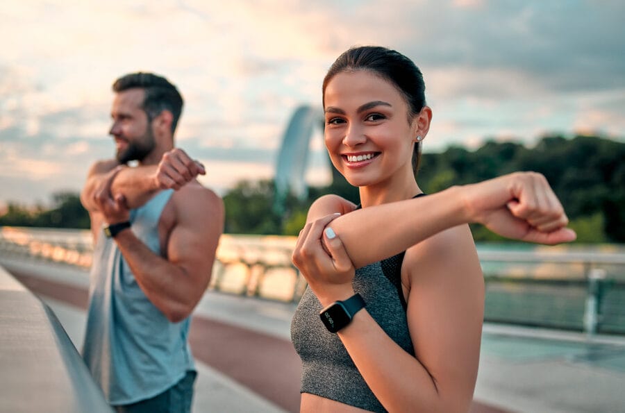 A smiling woman and man stretching their arms during an outdoor fitness session on a bridge at sunrise, demonstrating healthy posture - Corrective Exercise